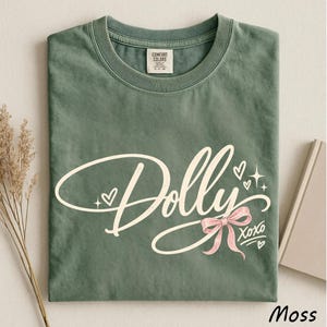 May include: A moss green crew neck t-shirt featuring the word Dolly in a large white cursive script font. The design includes small white hearts, four-pointed stars, a pink ribbon bow graphic, and the text XOXO in a white handwritten style font.