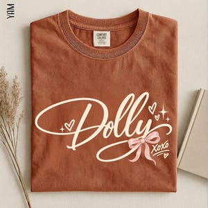 May include: A rust-colored short-sleeve t-shirt featuring the word Dolly in white cursive script. The design includes a light pink bow, small white hearts, and star accents, with the text XOXO printed in white near the bottom right corner.