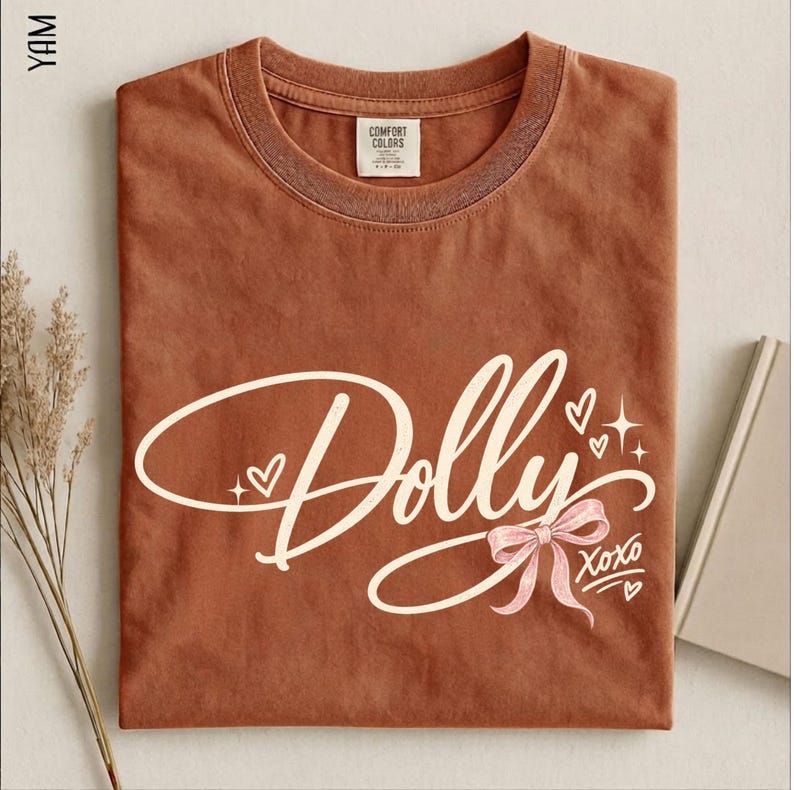 May include: A rust-colored short-sleeve t-shirt featuring the word Dolly in white cursive script. The design includes a light pink bow, small white hearts, and star accents, with the text XOXO printed in white near the bottom right corner.