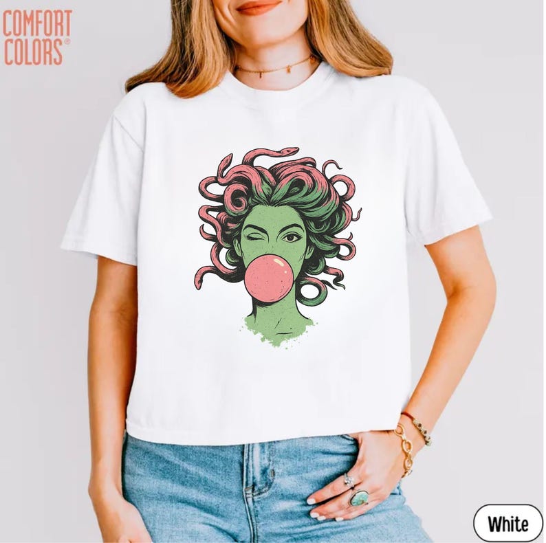 May include: White t-shirt featuring a cartoon illustration of a woman with green skin, pink hair, and a bubblegum bubble. The shirt has the words "Comfort Colors" in the upper left corner and "White" in the lower right corner.