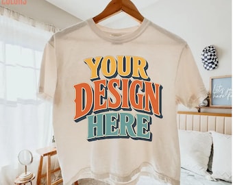 May include: An ivory short-sleeve t-shirt hanging on a wooden hanger. The shirt features the text "YOUR DESIGN HERE" printed in a bold, multi-layered font with yellow, orange, and teal colors. The fabric has a solid, light-colored appearance.