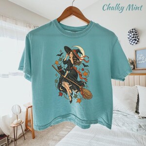 May include: A teal t-shirt featuring a graphic print of a person in a black dress riding a broomstick with a black cat, surrounded by autumn leaves, stars, bats, and a crescent moon. The shirt has a crew neckline and short sleeves.