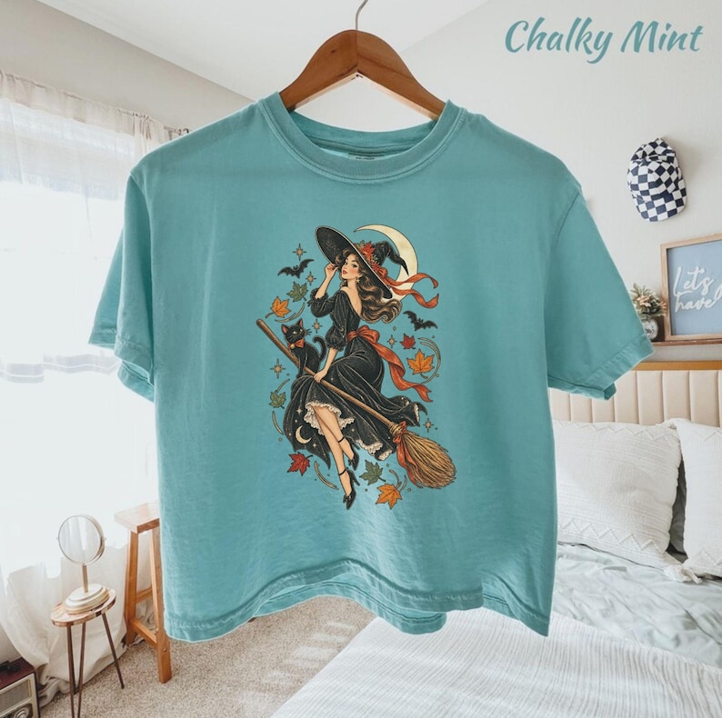 May include: A teal t-shirt featuring a graphic print of a person in a black dress riding a broomstick with a black cat, surrounded by autumn leaves, stars, bats, and a crescent moon. The shirt has a crew neckline and short sleeves.