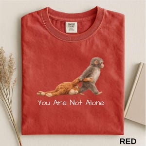 Puede incluir: Camiseta roja con un gr&aacute;fico de dos monos, uno tirando del otro. El texto "You Are Not Alone" est&aacute; impreso en blanco debajo de la imagen. La camiseta tiene cuello redondo y una etiqueta en la parte superior izquierda.