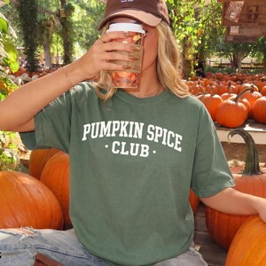 May include: A moss green short-sleeved t-shirt featuring the white text PUMPKIN SPICE CLUB printed in a collegiate font across the chest. The shirt is made of a solid-coloured fabric and has a crew neckline, providing a simple and casual appearance.