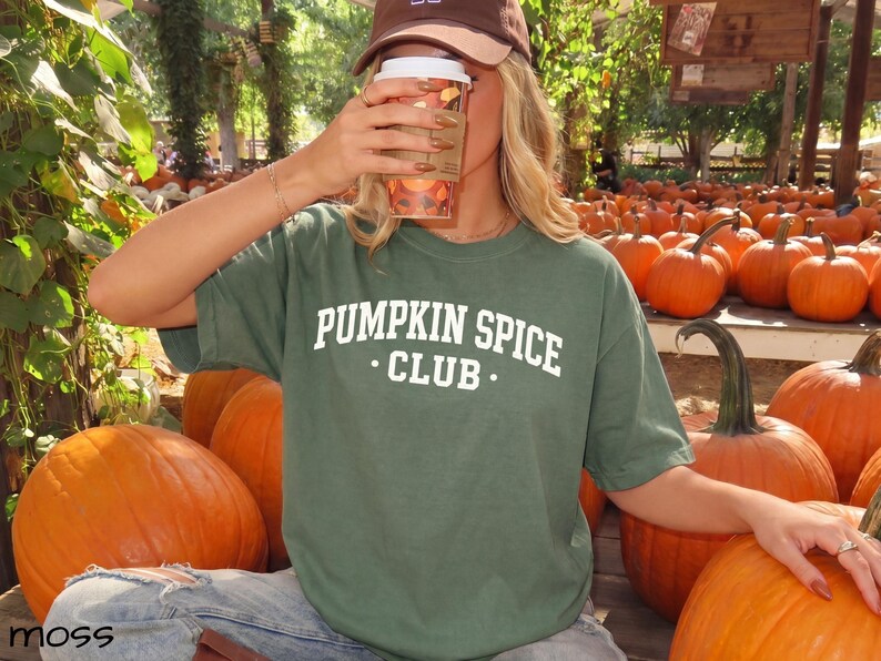 May include: A moss green short-sleeved t-shirt featuring the white text PUMPKIN SPICE CLUB printed in a collegiate font across the chest. The shirt is made of a solid-coloured fabric and has a crew neckline, providing a simple and casual appearance.