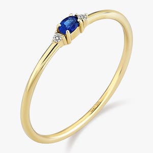 May include: A delicate gold ring featuring a vibrant blue oval-cut gemstone, flanked by two smaller white gemstones. The ring has a thin band and a polished finish, showcasing a minimalist design.