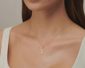 May include: A delicate gold-toned necklace featuring a small ankh pendant suspended from a thin, simple chain. The ankh symbol is centered on the collarbone area, providing a subtle and refined look that complements the light-colored neckline.