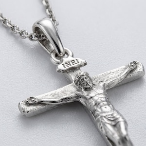 May include: A silver-toned metal crucifix pendant on a matching chain. The pendant features a detailed figure of a person on a cross with a textured surface. Above the figure, a small plaque is engraved with the letters INRI in capital letters.