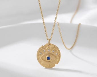 May include: A gold-toned necklace featuring a circular pendant with a textured, uneven edge. The pendant displays an embossed eye motif with a central blue stone and four small, clear accent stones arranged in an arc above the eye.