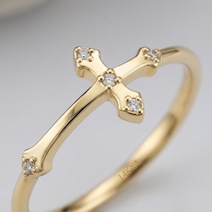 May include: A gold ring featuring a cross-shaped centerpiece adorned with five small, clear, round-cut stones. The band is smooth and polished, with the text 14K YA clearly engraved on the inner surface of the gold metal ring band.