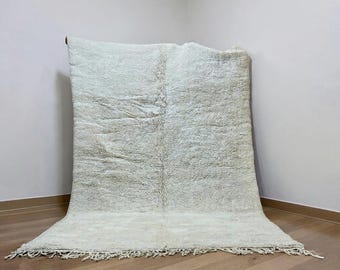 May include: A large, off-white rug with a shaggy texture. The rug is rectangular with a fringe along the bottom edge. The rug is displayed against a white wall and a wooden floor.