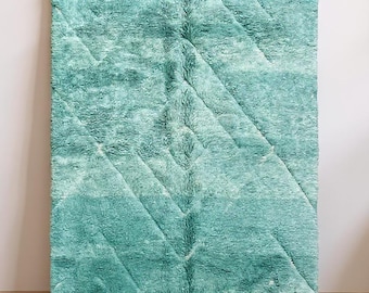 May include: A rectangular, turquoise rug with a geometric pattern of diagonal lines. The rug is hanging from a wooden rod. The rug has a shaggy texture and a fringe along the bottom edge.