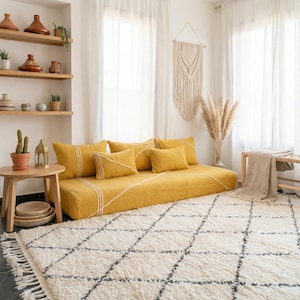 Op de afbeelding: Een gezellige woonkamer met een mosterdgele vloerbank en bijpassende kussens, en een crèmekleurig tapijt met een geometrisch patroon. Decoratieve planken, een houten bijzettafel en een macramé wandkleed maken de ruimte compleet.
