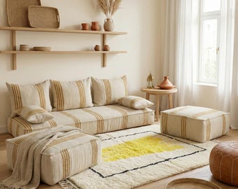Könnte beinhalten: Ein Wohnzimmer mit neutralen Farbtönen, das ein beiges Sofa und passende Ottomanen mit vertikalen Goldstreifen zeigt. Ein cremefarbener Teppich mit gelbem und schwarzem geometrischen Muster liegt auf dem Boden. Holzregale, ein Beistelltisch und ein geflochtener Tablett ergänzen die Einrichtung.