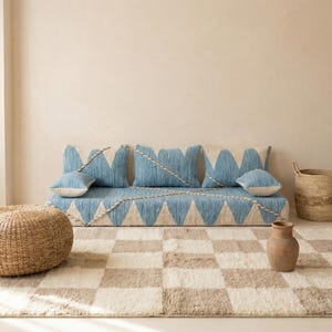 May include: A blue and cream-colored floor cushion with matching pillows and a rug. The cushion and pillows have a geometric design. A woven ottoman and basket are also in the image. The rug has a checkered pattern.