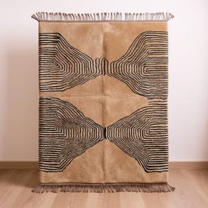 Puede incluir: Una alfombra rectangular beige con un diseño geométrico negro. La alfombra presenta un patrón repetitivo de líneas curvas, creando un diseño simétrico. La alfombra tiene flecos en los extremos cortos.