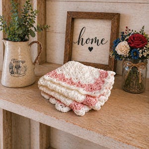 May include: A stack of white and pink crocheted coasters with a scalloped edge sits on a wooden shelf. A framed sign with the word "home" and a heart is in the background, along with a decorative pitcher and a jar of flowers.