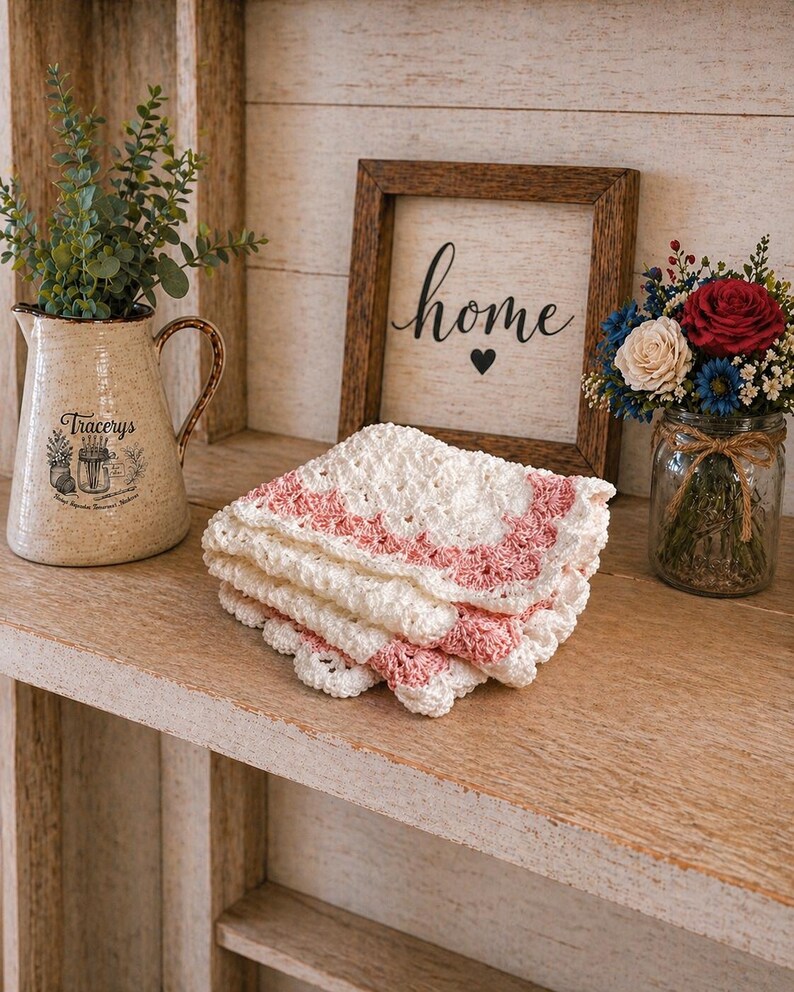 May include: A stack of white and pink crocheted coasters with a scalloped edge sits on a wooden shelf. A framed sign with the word "home" and a heart is in the background, along with a decorative pitcher and a jar of flowers.