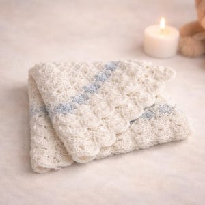 May include: A folded, off-white crocheted baby blanket with a light blue stripe detail. The blanket has a scalloped edge and is placed on a soft, light-colored surface. A lit candle and a teddy bear are in the background.