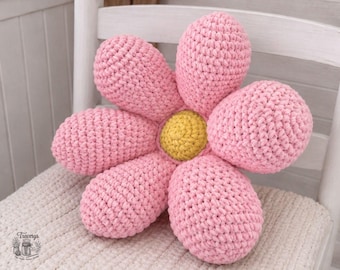 May include: A flower-shaped throw pillow made of chunky pink yarn with six rounded petals and a central yellow circular core. The crochet texture is visible throughout the surface of the pillow, which features a soft, plush appearance.