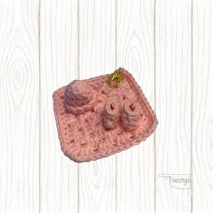 May include: A handmade pink crochet baby set, including a hat, booties, and a small square mat. A gold-colored decorative pin is attached to the mat. The set is designed for a baby.