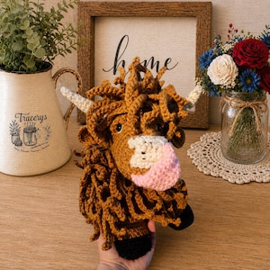 May include: A handmade crochet Highland cow head with brown yarn, white horns, and a pink and white muzzle. The cow has black hooves and is held in a person's hand. The background includes a framed sign that says "home".