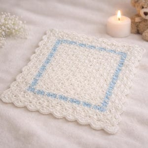 May include: A square, off-white crocheted doily with a light blue square border and scalloped edges. A lit white candle, a small teddy bear, and a sprig of white flowers are in the background.