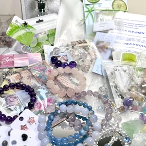 May include: A collection of beaded bracelets in various colors, including purple, pink, and blue, alongside small packets and cards. The bracelets appear to be made of gemstones and are displayed on a silver tray. Some packets have text.