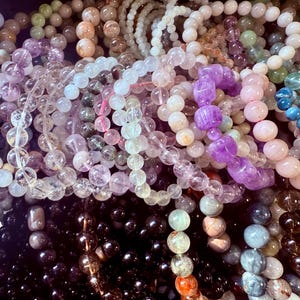 May include: A collection of beaded bracelets in various colors, including clear, purple, pink, green, and black. The beads are round and appear to be made of stone or crystal. Some bracelets have larger, carved beads.