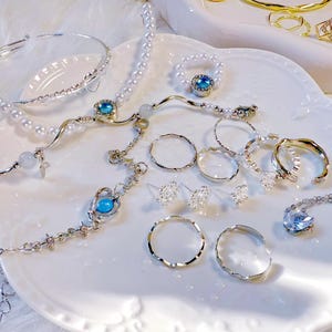 May include: A collection of silver-toned jewellery, including necklaces, bracelets, rings, and earrings. The pieces feature pearl and blue gemstone details. The jewellery is arranged on a white, scalloped-edge plate.