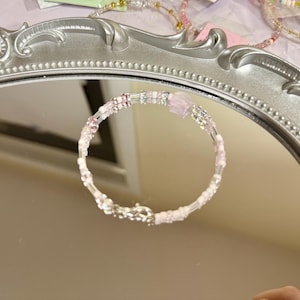 Puede incluir: Una delicada pulsera con una mezcla de cuentas transparentes y rosas, incluyendo una cuenta cuadrada rosa más grande. La pulsera se asegura con un cierre plateado. La joya se exhibe sobre un espejo con marco plateado.