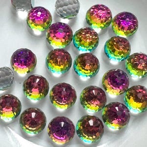 May include: A collection of iridescent, faceted glass beads in a white bowl. The beads display a spectrum of colors, including purple, green, and yellow. Three gray, faceted beads are also present. The beads are likely for jewelry making or crafting.