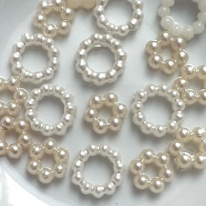 以下が含まれることがあります： 白とクリーム色の様々な色合いの、小さな花型のビーズのコレクション。 各ビーズは、より小さな丸いビーズで構成されており、繊細な花の模様を作り出しています。 ビーズは白いボウルの中に散らばっています。