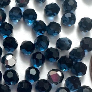 May include: A collection of faceted, dark blue crystal beads scattered on a white surface. The beads have multiple facets that reflect light, creating a sparkling effect. A copper penny is included for size reference.