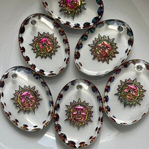 May include: Six clear, oval-shaped glass pendants with a sun design. The suns have a metallic, iridescent finish with pink, gold, and green hues. Each pendant has a small hole at the top for stringing. The edges are decorated with small, colorful dots.