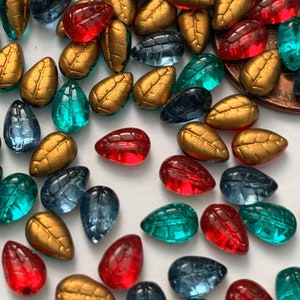 May include: Assortment of glass leaf-shaped beads in gold, red, teal, and blue colors. The beads are scattered on a white surface.