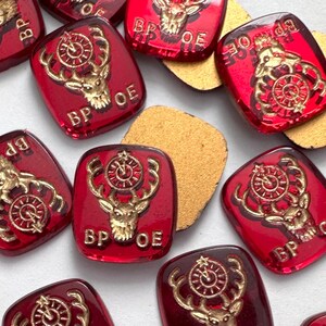May include: A collection of red, square-shaped glass cabochons with a gold-colored deer and clock design. Each cabochon has the letters "BP OE" imprinted on it. The cabochons are scattered on a white surface.