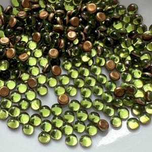May include: A collection of small, round beads in shades of green and bronze. The beads are scattered in a white bowl, with the green beads appearing translucent and the bronze beads opaque. The beads are likely used for jewelry making or crafting.