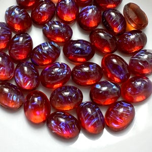May include: A collection of 25 oval-shaped iridescent glass cabochons in shades of red and purple. The cabochons have a smooth, polished surface and a variety of swirling patterns.