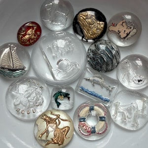 May include: A collection of clear glass cabochons with various designs, including a sailboat, a mermaid, a fish, and a life preserver. Some cabochons have gold or silver accents. The cabochons are in a white bowl.