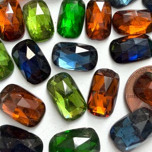 May include: A collection of faceted, rectangular gemstones in various colors, including green, orange, blue, and brown. The gemstones are arranged on a white surface, with a penny included for size reference. The gems have a polished, reflective surface.