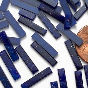 May include: A collection of small, rectangular, blue glass tiles with gold flecks. The tiles are scattered on a white surface, with a US penny for scale.