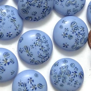 May include: A collection of light blue, round beads with a flat back and a hole drilled through the center. Each bead is decorated with a pattern of small, stylized flowers and striped orbs in shades of blue and white. The beads are scattered across a white surface.