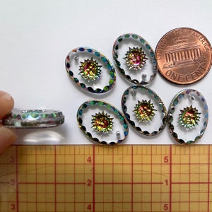 May include: Oval-shaped clear glass beads with a sunburst design in the center. The sunburst has iridescent colors. The beads are approximately 1 inch in length. A U.S. penny is included for size comparison. The beads have a scalloped edge.
