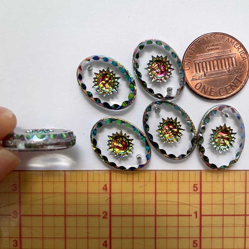 May include: Oval-shaped clear glass beads with a sunburst design in the center. The sunburst has iridescent colors. The beads are approximately 1 inch in length. A U.S. penny is included for size comparison. The beads have a scalloped edge.