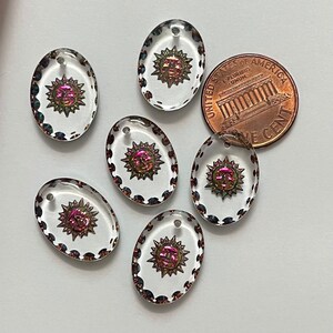 May include: Six clear, oval-shaped pendants with a sun design in the center. The sun has a face and is surrounded by rays. The pendants have a decorative border and a small hole at the top. A US penny is next to the pendants.