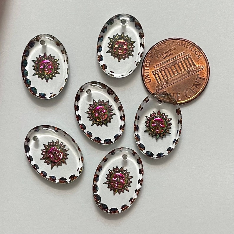May include: Six clear, oval-shaped pendants with a sun design in the center. The sun has a face and is surrounded by rays. The pendants have a decorative border and a small hole at the top. A US penny is next to the pendants.
