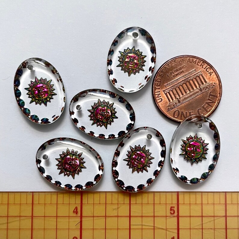 May include: Six clear oval-shaped glass pendants with a sunburst design in the center. The sunburst is a mix of pink, green, and gold. Each pendant has a small hole at the top for stringing. A penny is included for size reference.