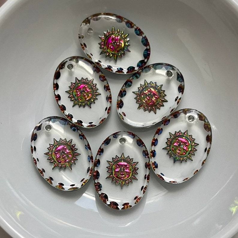 May include: Oval-shaped clear glass pendants with a sun design. The sun has a face and radiating rays with iridescent colors. The edges of the pendants are decorated with small, colorful beads. Each pendant has a small hole at the top.